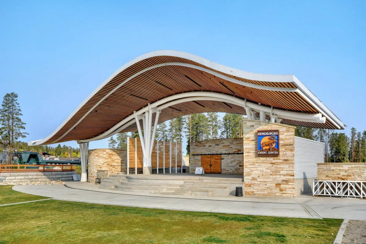 This lively exterior scene features the Rendezvous Event Center at Hideaway Park, showcasing a curved roof structure, a welcoming stone façade, and an open stage area set up for outdoor concerts and community gatherings. The festive mood is enhanced by an audience area ready for performances and celebratory events under the stars.