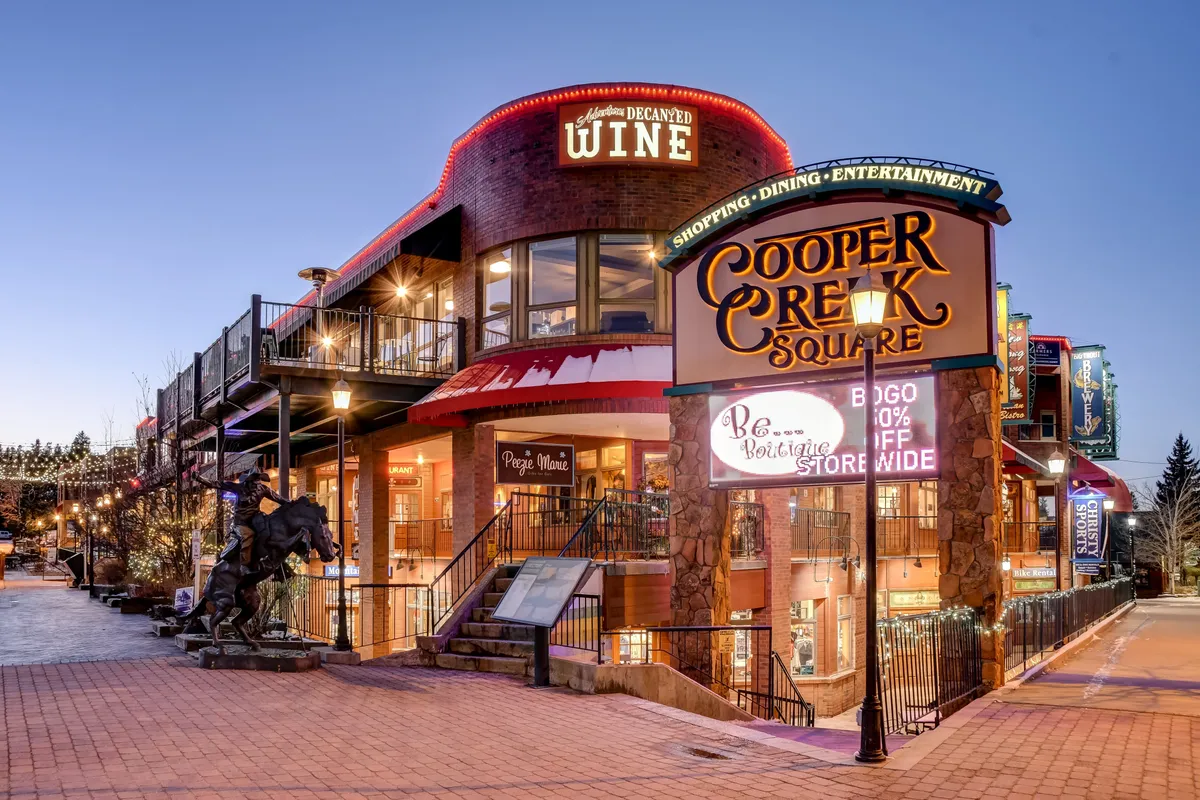 A bustling exterior scene highlighting Cooper Creek Square, with its prominent signage, wine shop, decorative lighting, and a staircase leading up to a vibrant plaza. The area is clearly a hub for shopping, dining, and social gatherings, with an inviting and festive atmosphere.