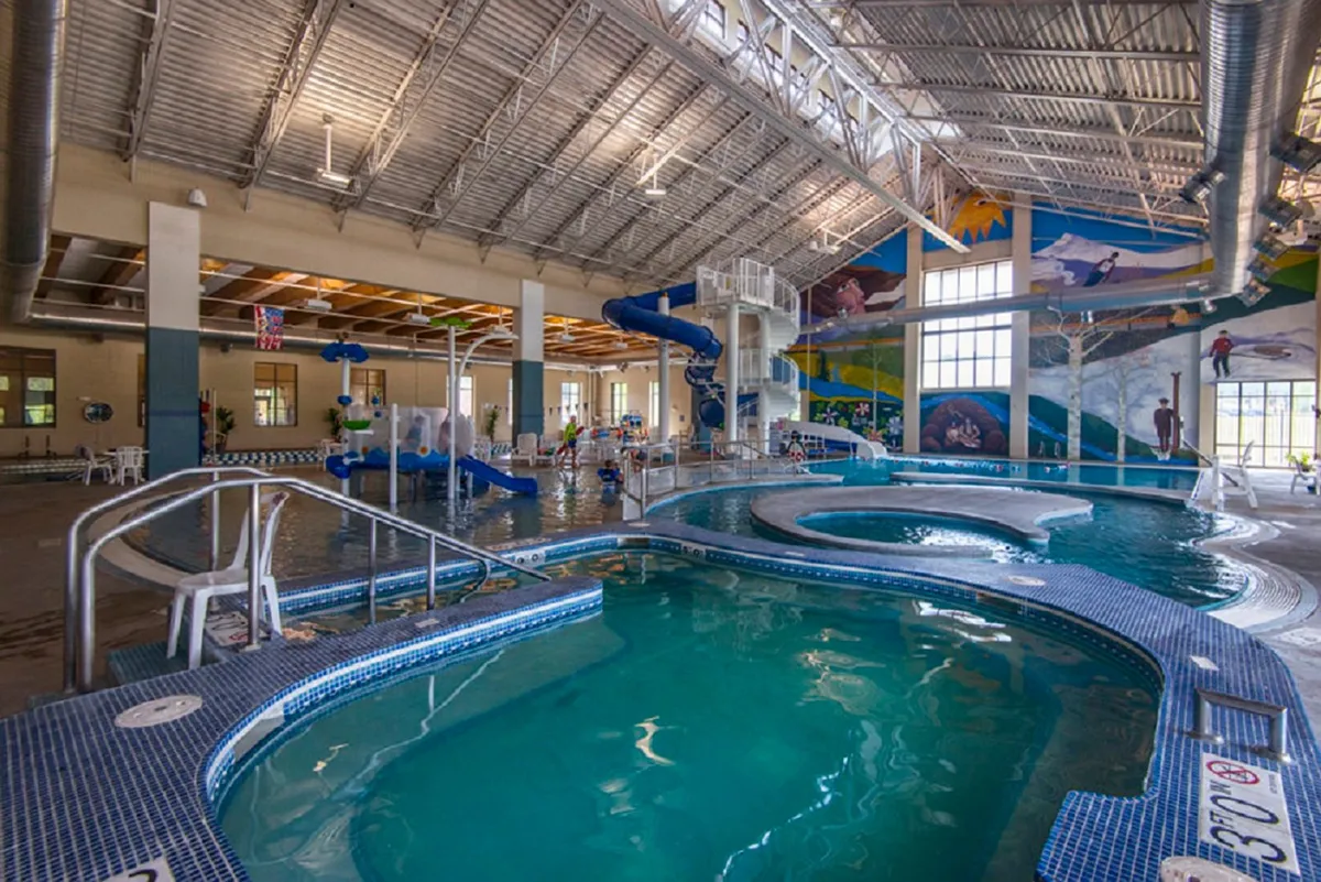 A lively interior scene of a recreation center pool area, with water slides, colorful murals, and children playing in the water. The atmosphere is energetic and family-friendly, showcasing amenities for all ages.