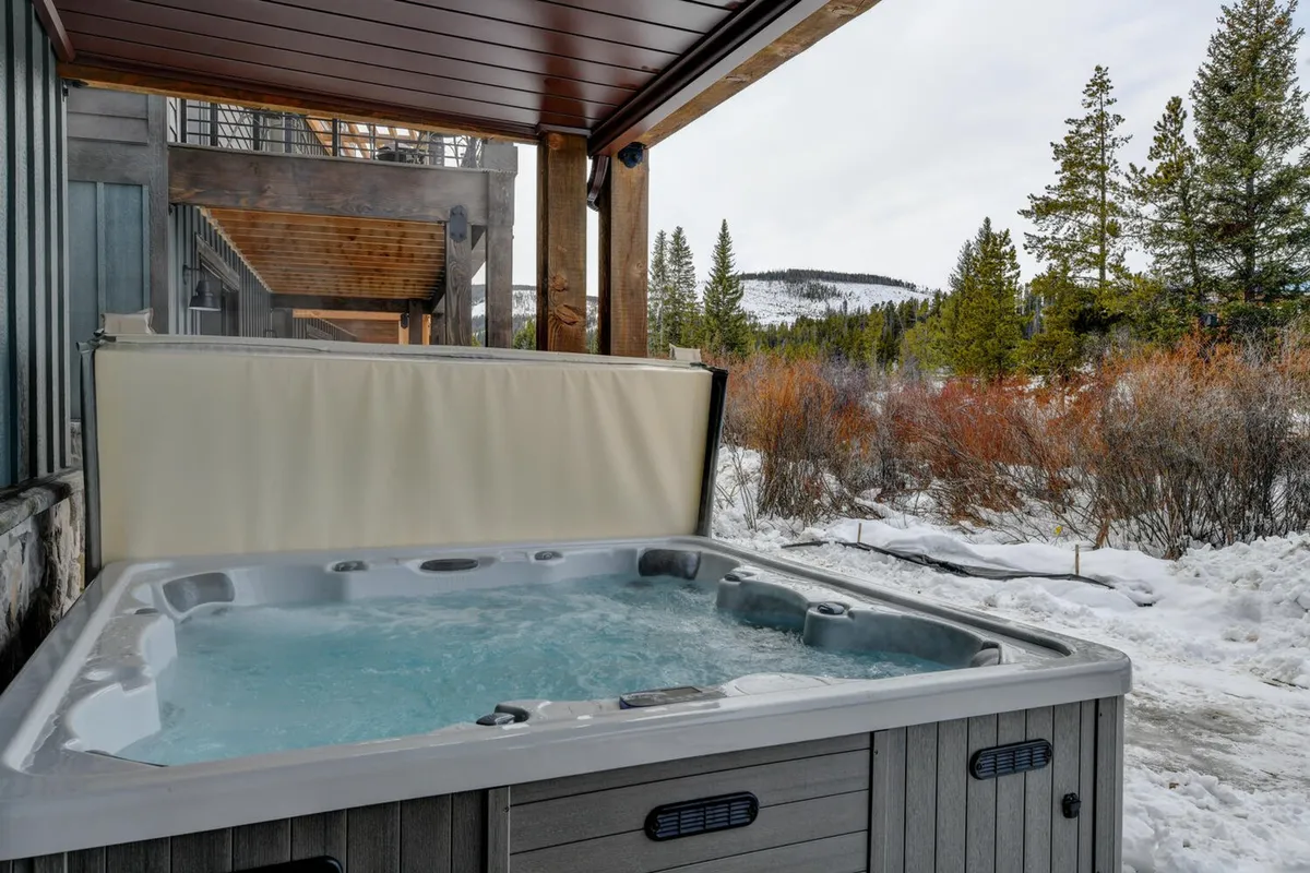 A serene exterior view of a private hot tub set against a snowy landscape, with a wooden structure providing shelter and a backdrop of gentle mountain slopes. The scene evokes tranquility, highlighting the opportunity to soak while surrounded by natural beauty, with sunset hues visible over the mountains.