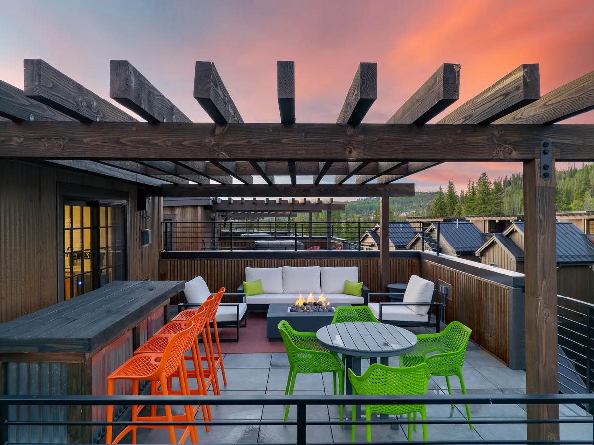 An inviting exterior lounge area featuring a vibrant mix of colorful chairs arranged around a modern fire pit, all beneath a wooden pergola. The setting overlooks natural landscape views, creating a seamless connection between the comfort of the home and the outdoors. The space is designed for socializing, relaxation, and enjoying both day and evening activities.