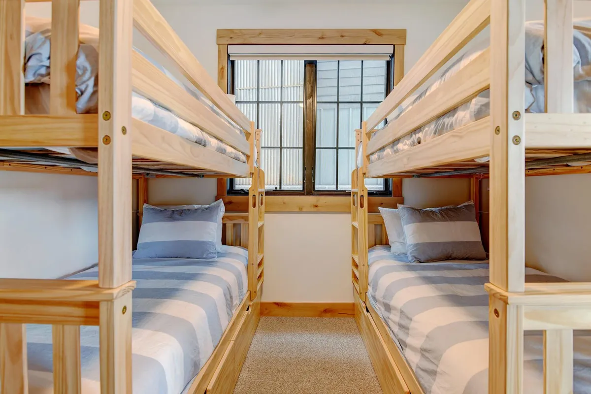 A bright and cozy bunk room with two sets of bunk beds, striped bedding, and a window that lets in natural light. The communal arrangement invites togetherness, and the space feels playful and perfect for kids or group sleepovers.