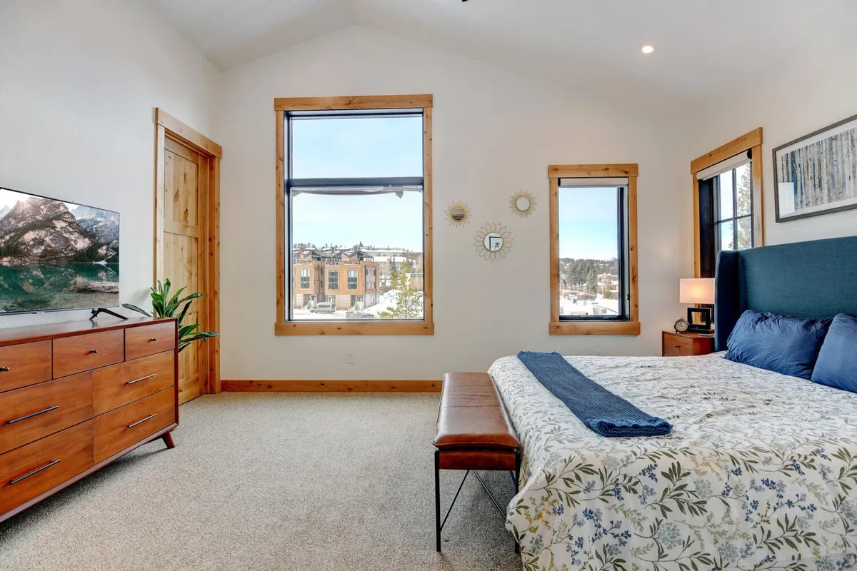 A serene bedroom with large windows framing outdoor views, a comfortable bed with floral bedding, a wooden dresser, and decorative mirrors. The space exudes tranquility and features a television for added relaxation, perfectly illustrating a restful private retreat.