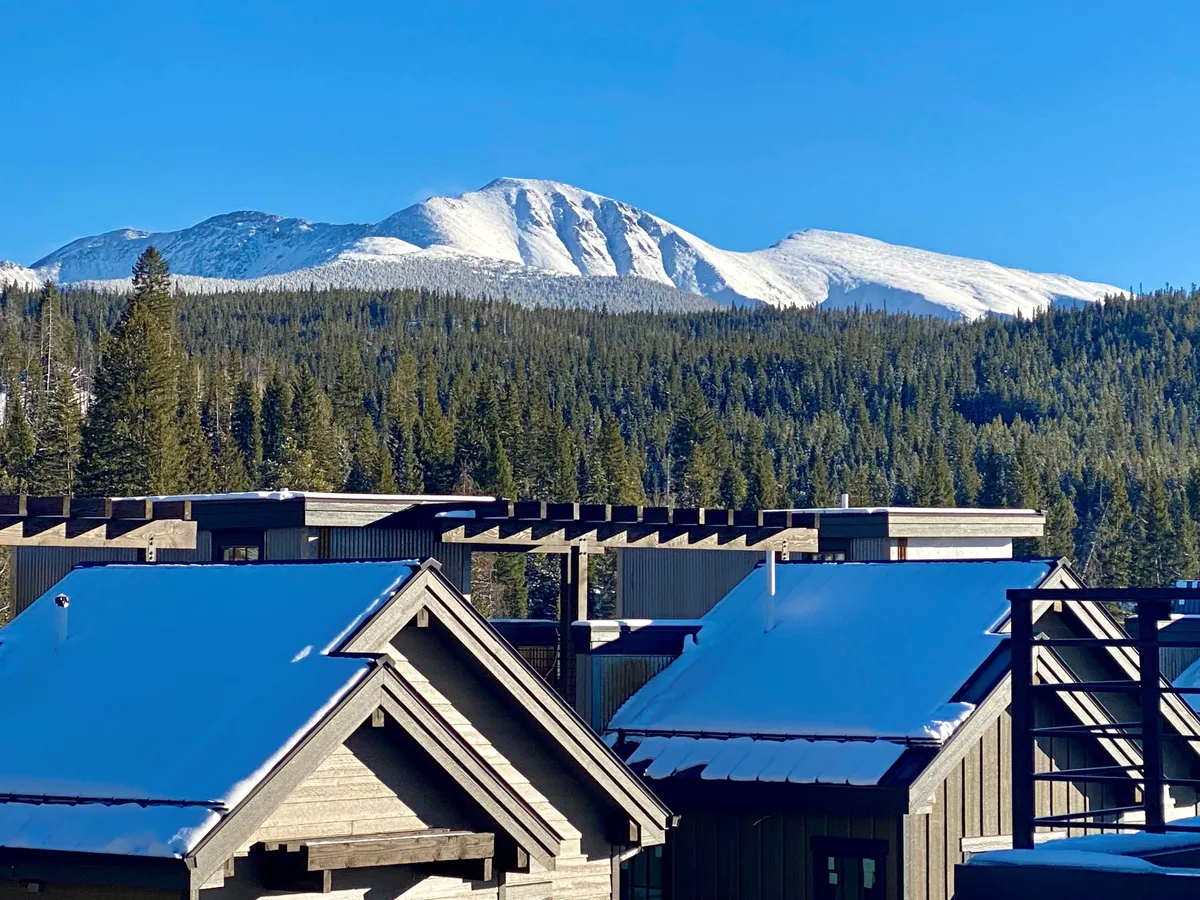 A panoramic view of snow-capped mountains and dense pine forests, with modern building rooftops dusted in snow. The scene hints at nearby ski slopes and opportunities for hiking and outdoor photography, set against a serene alpine backdrop.