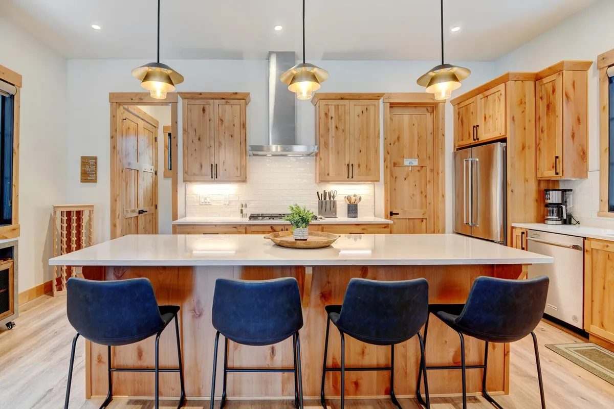 A modern chef’s kitchen with a central island, wooden cabinetry, stainless steel appliances, and pendant lighting. The layout invites interaction, with ample space for cooking and socializing, and a clear view into the gathering area.