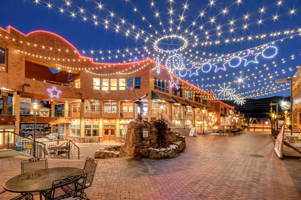 An evening exterior shot of a lively downtown area, featuring string lights, decorative stars, a cobblestone path, comfortable seating, and a rock fountain. The space is designed for relaxed dining, socializing, strolling, and festive community events, radiating a cheerful and inviting mood.