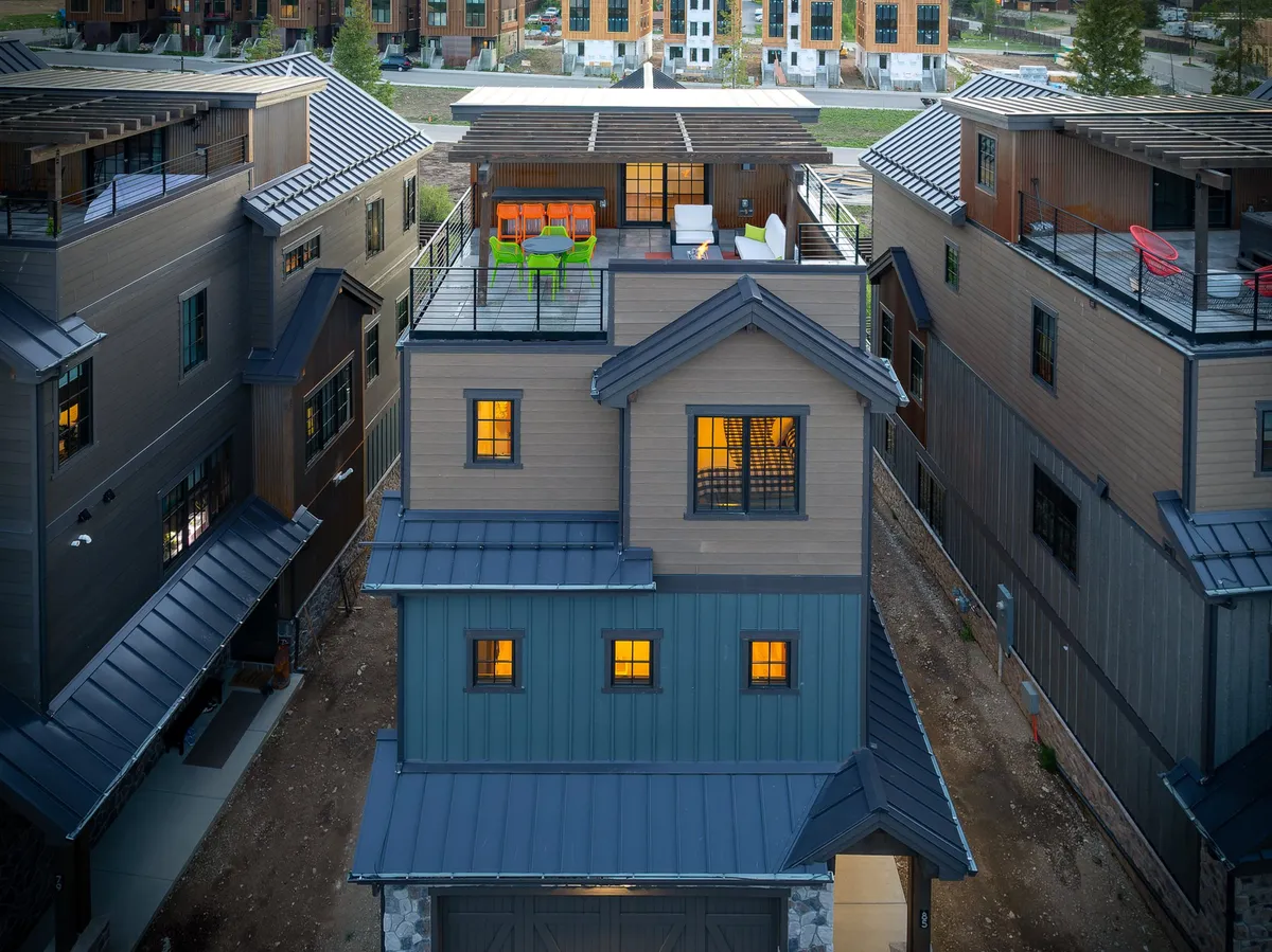 An elevated exterior shot of a rooftop terrace at dusk, featuring illuminated windows, colorful lounge furniture, and sweeping views of the landscape. The inviting setup includes cozy seating and a sense of warmth, suggesting a perfect spot for sunset cocktails and fireside conversations.