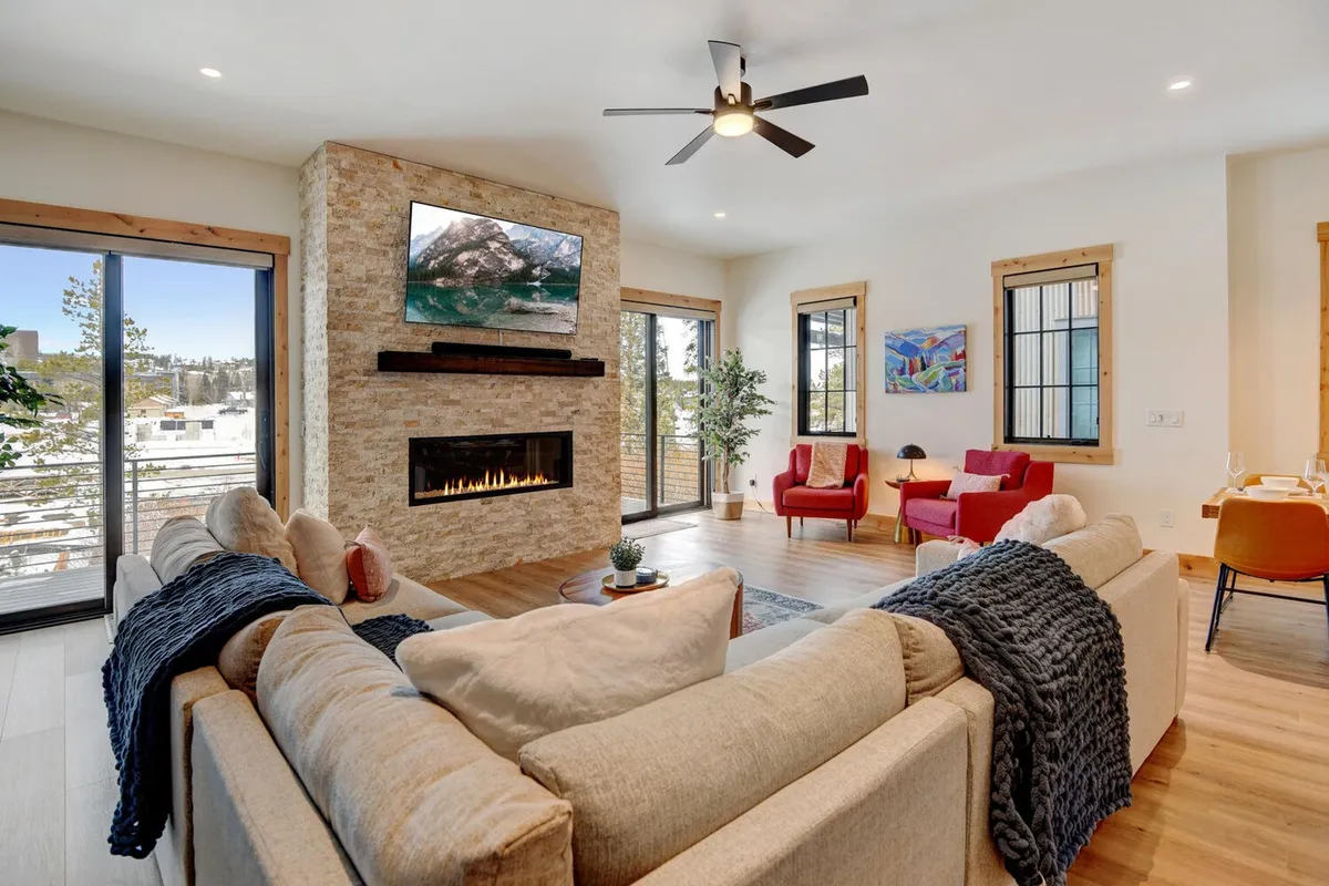 A welcoming living room featuring a plush sofa, red accent chairs, a sleek fireplace, large windows that flood the space with natural light, and contemporary artwork. The space appears warm, social, and ideal for gathering, with a large TV above the fireplace.