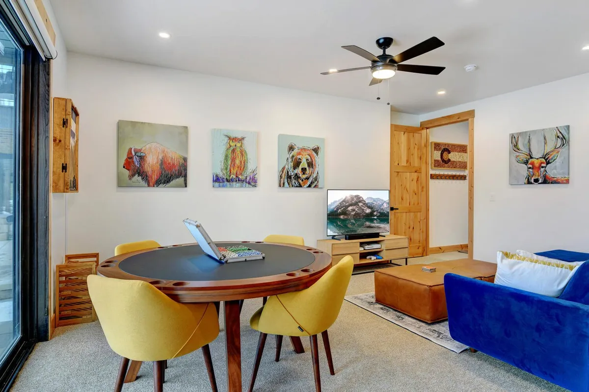 A cheerful dining and living space featuring a large table with yellow chairs, colorful animal artwork, a blue sofa, and a television with a scenic view. The decor is playful, welcoming to kids, and offers comfort for adults and pets alike.