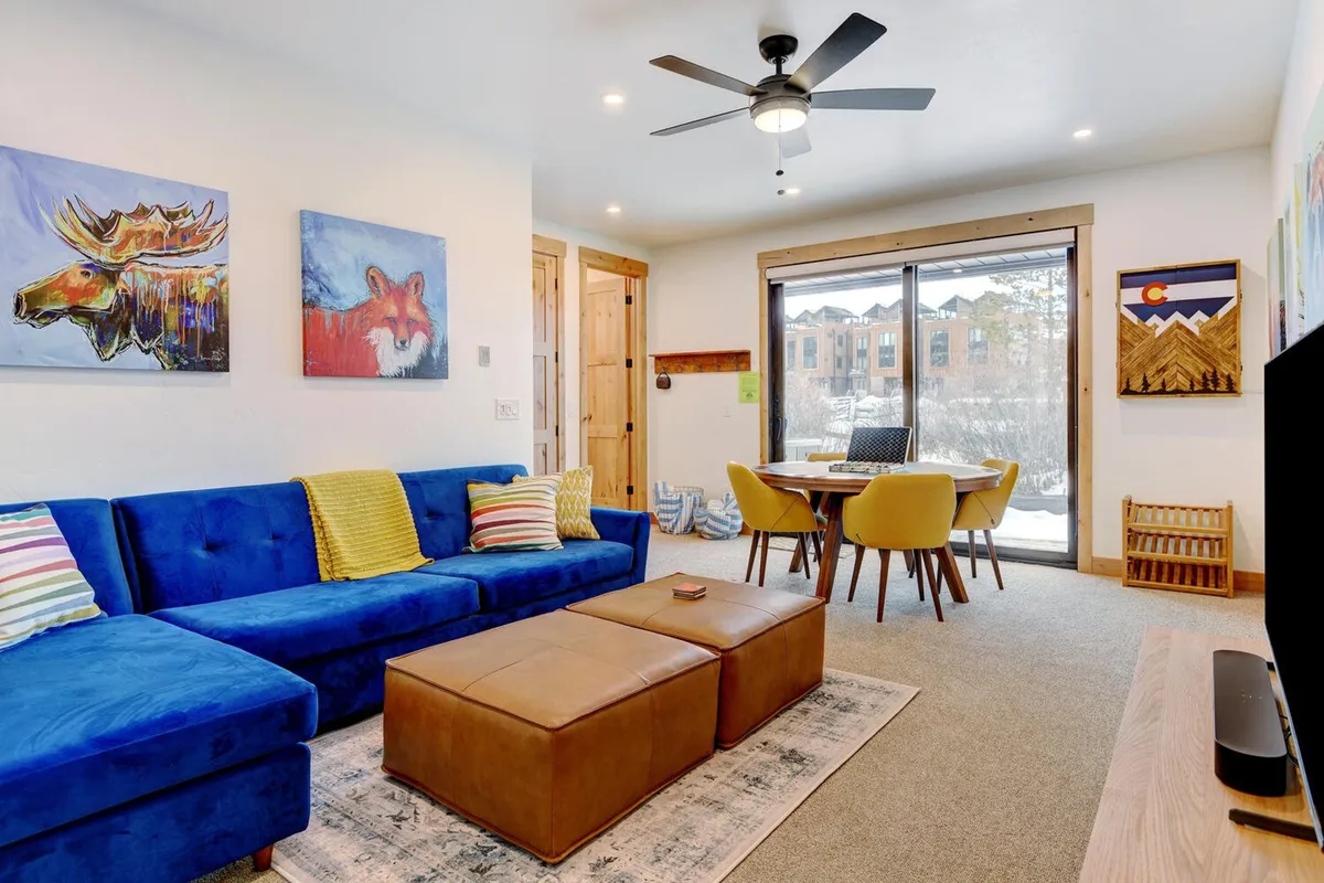 A spacious living area with a blue sectional sofa, colorful artwork, a wooden dining table, yellow chairs, and large windows. The setting is ideal for group activities, board games, and family relaxation, with plenty of seating for all ages.