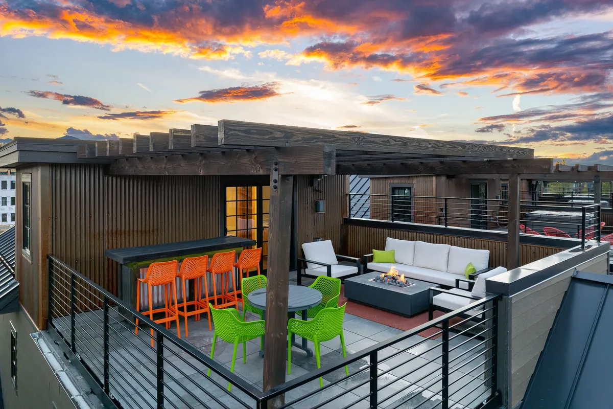 The image showcases the villa's inviting outdoor lounge area, featuring a fire pit surrounded by colorful chairs, a sleek bar counter with stools, and ample space for socializing. The scene is set against the backdrop of a mountain skyline at sunset, creating a festive yet cozy atmosphere ideal for gatherings.